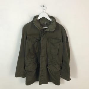 May include: An olive green military-style jacket with multiple front pockets and a high collar. The jacket is suspended on a white hanger against a white backdrop. The jacket has a patch on the upper arm.