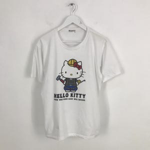 May include: White t-shirt featuring a cartoon image of Hello Kitty wearing a hard hat and holding a hammer and a tape measure. The text "Hello Kitty Those who work hard will succeed." is printed below the image.