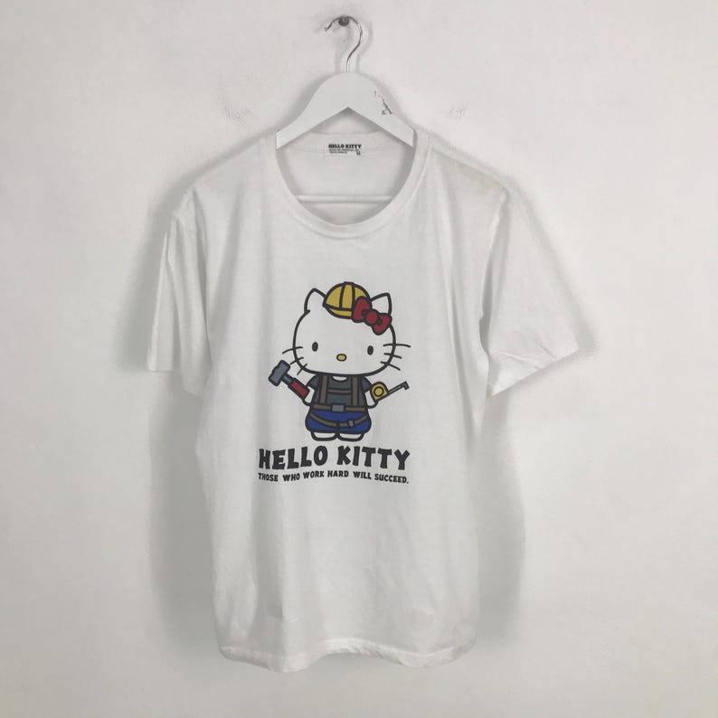 May include: White t-shirt featuring a cartoon image of Hello Kitty wearing a hard hat and holding a hammer and a tape measure. The text "Hello Kitty Those who work hard will succeed." is printed below the image.