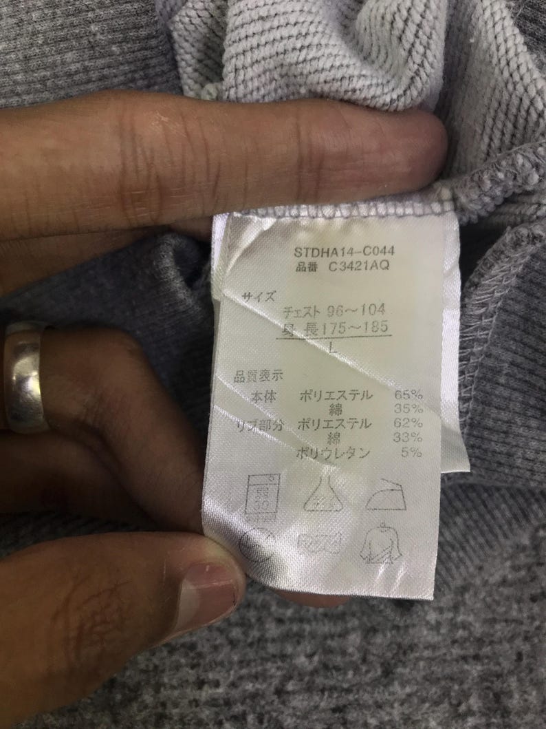 May include: A close-up of a grey sweatshirt label with text. The label includes the product code STDHA14-C044 and size information. The label also lists the fabric composition: 65% polyester, 35% cotton, 62% polyester, 33% cotton, and 5% polyurethane.