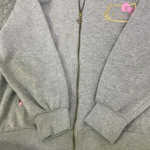 May include: A gray zip-up hoodie featuring a gold-outlined Hello Kitty design with a pink bow on the upper left chest. The hoodie has a full-length zipper and ribbed cuffs and hem. The sleeves are long and the fabric appears to be a soft, comfortable material.