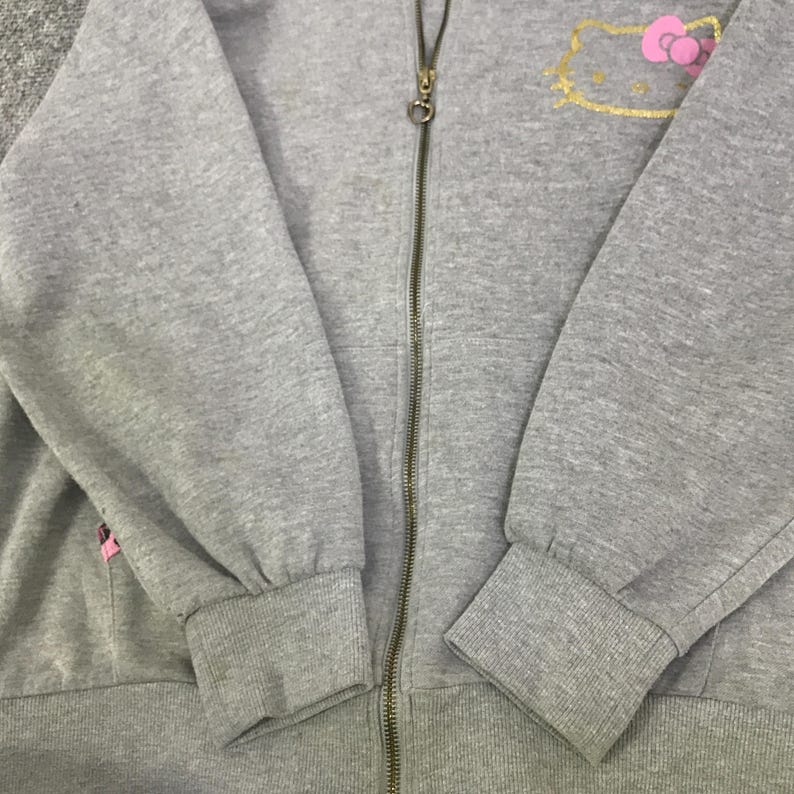 May include: A gray zip-up hoodie featuring a gold-outlined Hello Kitty design with a pink bow on the upper left chest. The hoodie has a full-length zipper and ribbed cuffs and hem. The sleeves are long and the fabric appears to be a soft, comfortable material.