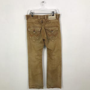 May include: A pair of brown denim jeans with a distressed look. The jeans have a straight leg and a relaxed fit. The back pockets have a unique design with orange stitching. The jeans have a label that says "EDWIN" on the back.