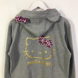 May include: A gray zip-up hoodie featuring a gold Hello Kitty design on the back. The hoodie has a pink and black leopard print bow and ear accents. The words "Hello Kitty" are printed in gold below the character.