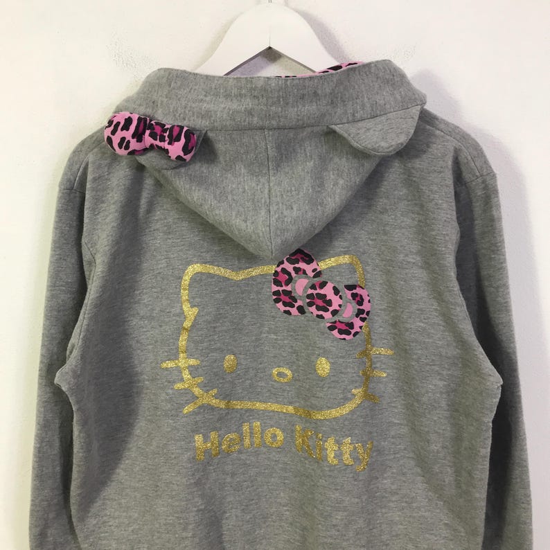 May include: A gray zip-up hoodie featuring a gold Hello Kitty design on the back. The hoodie has a pink and black leopard print bow and ear accents. The words "Hello Kitty" are printed in gold below the character.