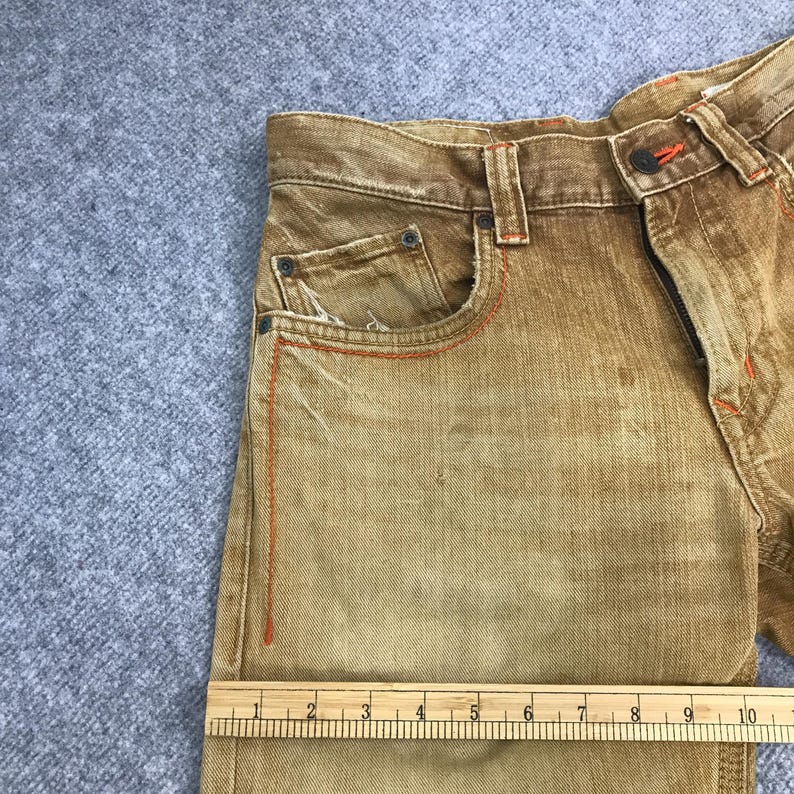 May include: Close-up of a pair of brown denim jeans with orange stitching. The jeans have a front pocket and a ruler is placed on the bottom of the image to show the size.
