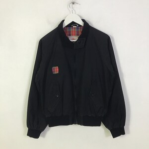 以下が含まれることがあります： リブ編みの襟、袖口、裾が特徴の黒いジップアップジャケット。内側の裏地には赤、緑、青のチェック柄が施されています。右胸には小さな四角いチェック柄のパッチがあり、ボタン付きのサイドポケットが2つ付いています。