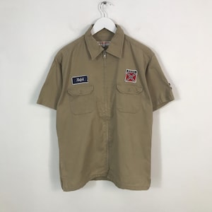 Puede incluir: Camisa de trabajo caqui de manga corta con cremallera frontal y dos bolsillos en el pecho. Cuenta con un pequeño parche que dice "Ralph" y un parche de la bandera confederada. La camisa está colgada de una percha blanca.