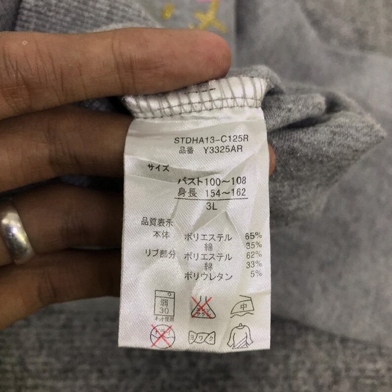 May include: Close-up of a gray garment tag with Japanese text. The tag includes details like "STDHA13-C125R" and fabric composition percentages. The tag is held by a hand, with a silver ring visible.