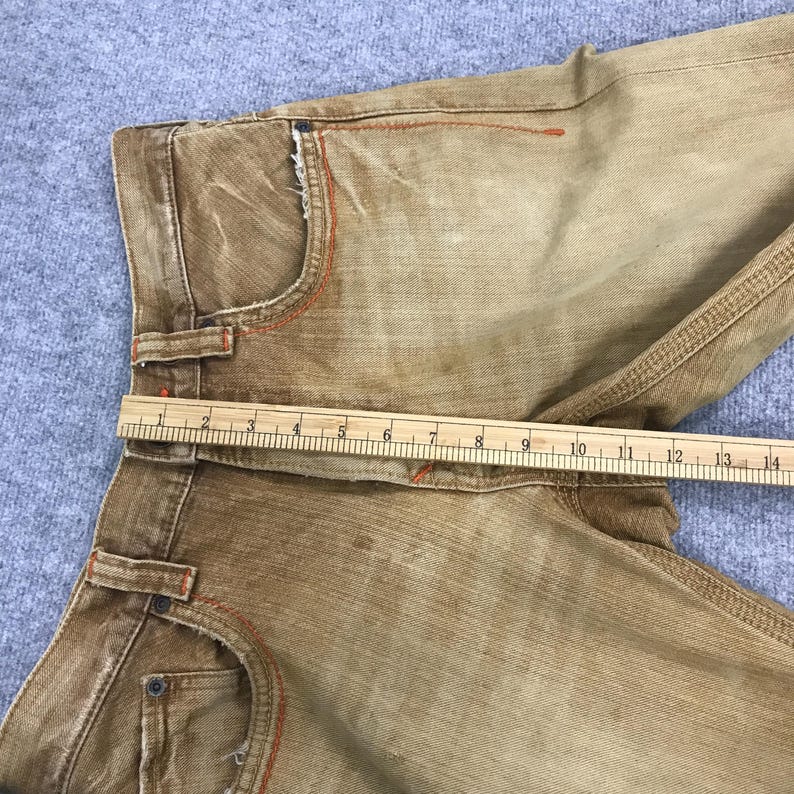 May include: A pair of brown denim jeans with orange stitching. The jeans are laid flat and a ruler is placed across the waistband. The ruler measures 35.5 cm.
