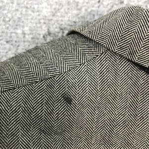 May include: Close-up of a black and white herringbone pattern fabric. The fabric is tightly woven with a repeating V-shaped design. The image shows the texture and detail of the material, with a small dark spot visible.