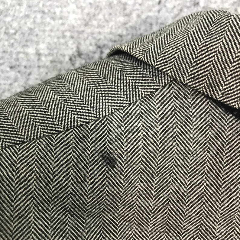 May include: Close-up of a black and white herringbone pattern fabric. The fabric is tightly woven with a repeating V-shaped design. The image shows the texture and detail of the material, with a small dark spot visible.