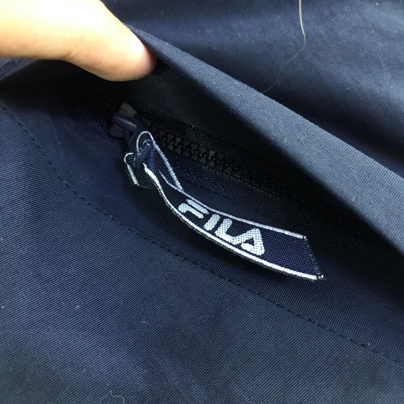May include: Close-up of a navy blue jacket zipper with a white and navy blue FILA logo tag. The zipper is partially open, revealing the interior lining of the jacket. The fabric appears to be a smooth, durable material.