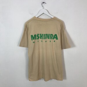 May include: Tan t-shirt with the text "MSHINDA WINNER" in green block letters. The shirt is hanging on a white hanger against a white background. The t-shirt has short sleeves and a crew neck.