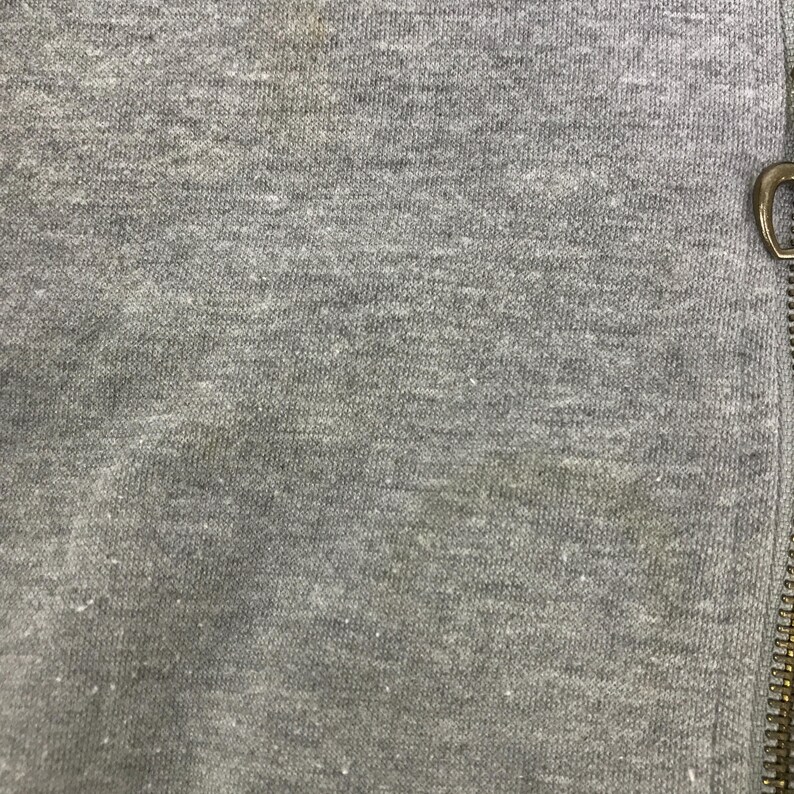 May include: Close-up of a heather gray fabric with a visible texture. A gold-colored zipper is on the right side of the image. The fabric appears to be a soft knit material, possibly from a sweatshirt or similar garment.