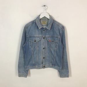 May include: Light blue denim jacket with a button-down front, two chest pockets, and two side pockets. The jacket has a classic collar and a small orange logo on the chest pocket. The jacket is hanging on a white hanger.