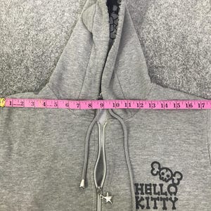 May include: Grey zip-up hoodie with a black and white patterned hood lining. The hoodie has a silver zipper with a star-shaped charm and the words "HELLO KITTY" with a skull and crossbones graphic.