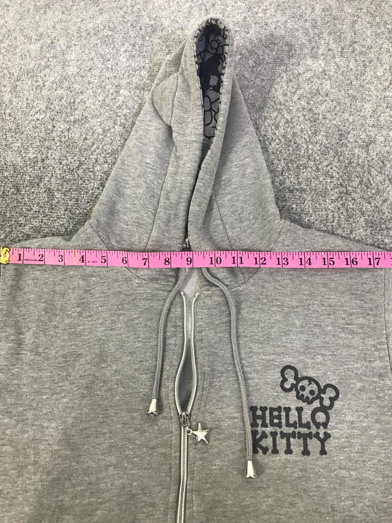 May include: Grey zip-up hoodie with a black and white patterned hood lining. The hoodie has a silver zipper with a star-shaped charm and the words "HELLO KITTY" with a skull and crossbones graphic.