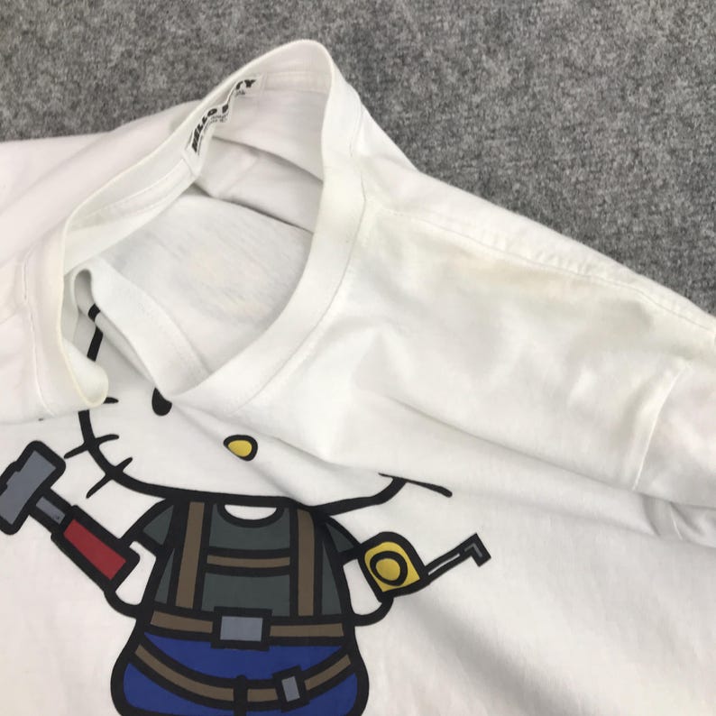 May include: White t-shirt featuring a cartoon illustration of Hello Kitty dressed as a construction worker holding a hammer. The text "Hello Kitty" is printed on the collar.