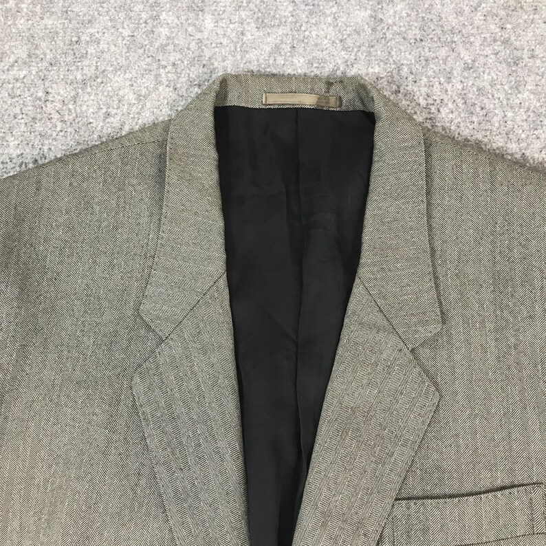 May include: A close-up of a grey blazer with a black interior lining. The blazer features a notched lapel and a single pocket. The fabric has a subtle textured pattern.