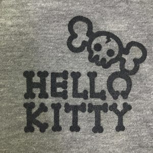 May include: Grey t-shirt featuring a black skull and crossbones design above the words "HELLO KITTY". The text is in a bold font, and the design is centrally placed.