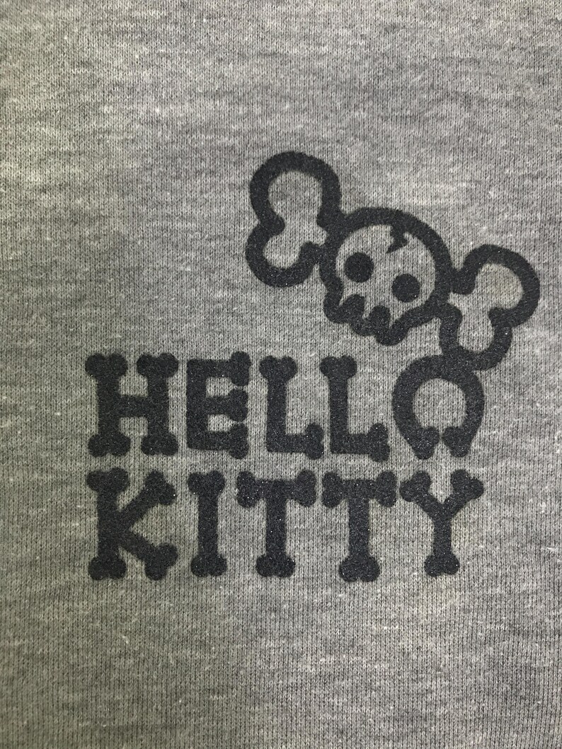 May include: Grey t-shirt featuring a black skull and crossbones design above the words "HELLO KITTY". The text is in a bold font, and the design is centrally placed.