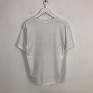 May include: A plain white t-shirt hanging on a white hanger against a white wall.
