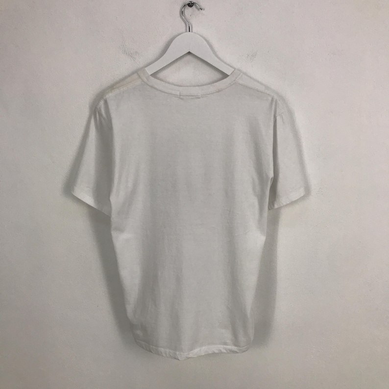 May include: A plain white t-shirt hanging on a white hanger against a white wall.