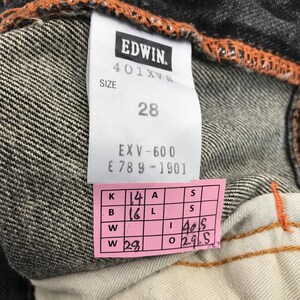 May include: Close-up of a denim tag from Edwin jeans. The tag displays the brand name, size 28, and other details. A pink tag with measurements is also visible. The jeans have orange stitching.