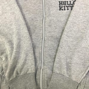 May include: A grey zip-up hoodie with a hood and a white zip. The hoodie features the text "HELLO KITTY" with a skull and crossbones graphic above it. The cuffs and waistband are ribbed.