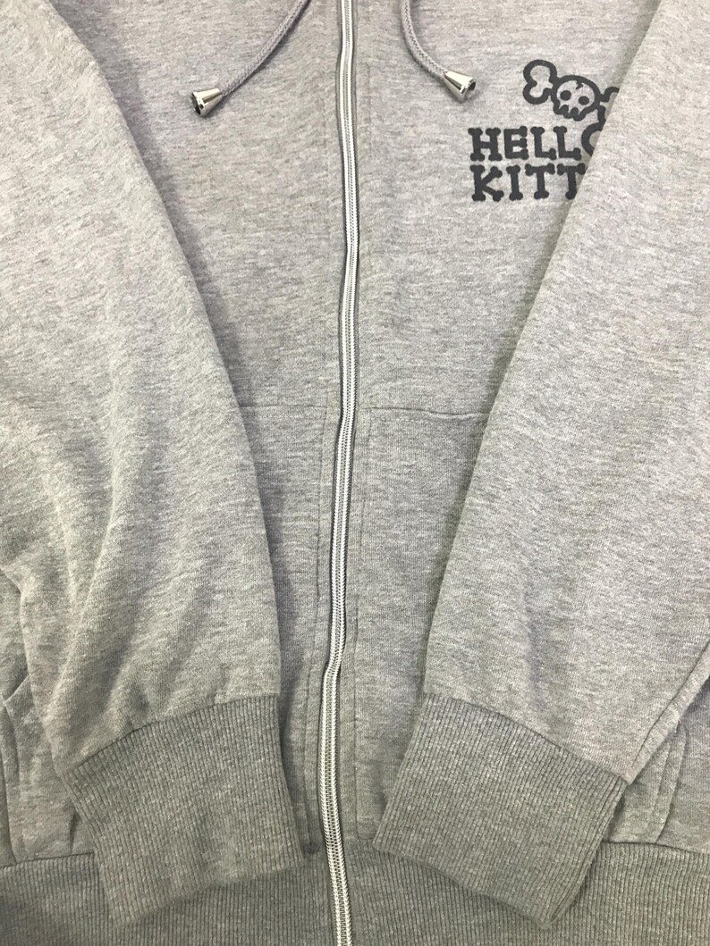 May include: A grey zip-up hoodie with a hood and a white zip. The hoodie features the text "HELLO KITTY" with a skull and crossbones graphic above it. The cuffs and waistband are ribbed.
