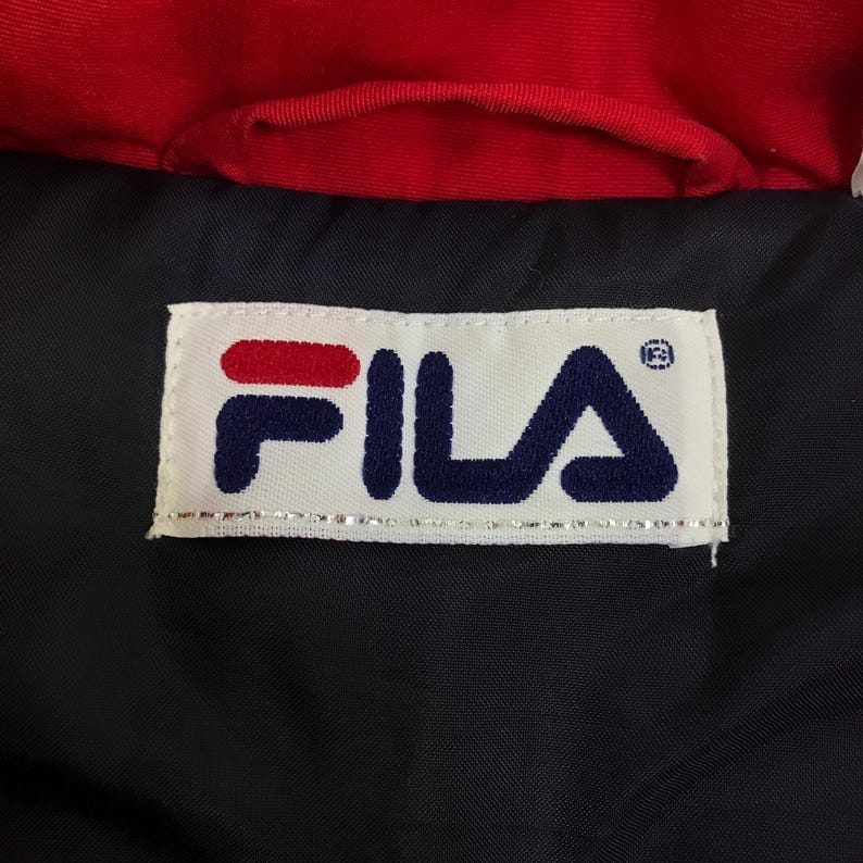May include: Close-up of a black garment with a red collar. A white rectangular label with the brand name "FILA" in navy blue and red is sewn onto the garment. The label has a silver border.