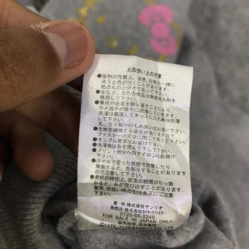 May include: Close-up of a gray fabric tag with Japanese text and the words "FOR SALE IN JAPAN ONLY." The tag also includes the Sanrio copyright information. The tag is attached to a gray garment.