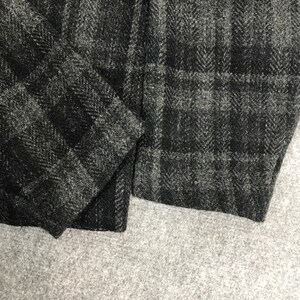 May include: Close-up of a section of dark grey and black tweed fabric. The fabric has a subtle herringbone weave and a plaid pattern. The material appears to be thick and textured, with a slightly rough appearance.