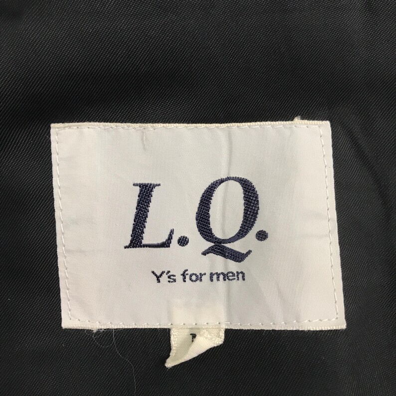 May include: A close-up shot of a white label with the text "L.Q." in dark blue, and "Y's for men" below. The label is attached to a dark fabric, possibly a piece of clothing.