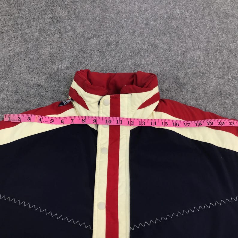 May include: A close-up of a navy blue and red jacket with a white collar and accents. The jacket has a red stripe down the center and a zigzag pattern along the bottom. A measuring tape is laid across the jacket.