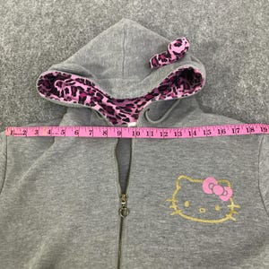 May include: A gray zip-up hoodie with a Hello Kitty design. The hood lining and a small bow on the design are pink leopard print. The Hello Kitty face is gold, and the bow is pink. The zipper has a heart-shaped pull.