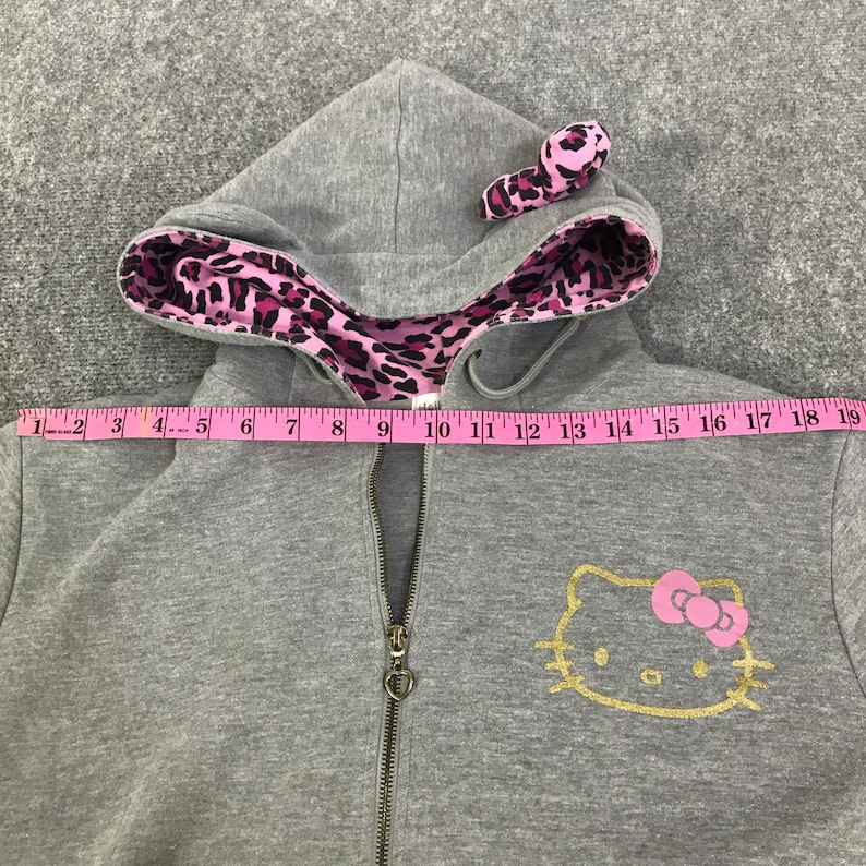 May include: A gray zip-up hoodie with a Hello Kitty design. The hood lining and a small bow on the design are pink leopard print. The Hello Kitty face is gold, and the bow is pink. The zipper has a heart-shaped pull.