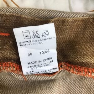 May include: A brown cotton label with white text and a washing instruction symbol. The label says "100% Cotton" and "Made in China". The label also includes a phone number and a warning about colour fading.