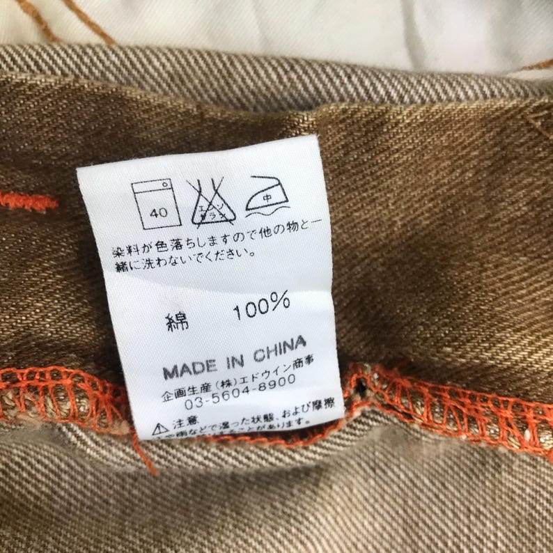 May include: A brown cotton label with white text and a washing instruction symbol. The label says "100% Cotton" and "Made in China". The label also includes a phone number and a warning about colour fading.