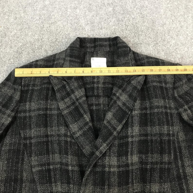 May include: A grey and black plaid blazer with a notched collar. A wooden ruler is placed across the blazer, measuring its width. The blazer has a herringbone pattern and a tag with the letter "Y".