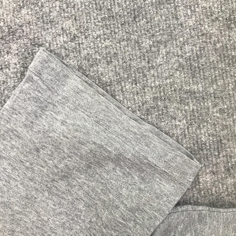 Pu&ograve; includere: Primo piano di un tessuto grigio, possibilmente una manica o un orlo di un capo. Il tessuto ha un aspetto strutturato, con dettagli visibili di maglia o tessitura. Il bordo &egrave; ripiegato e cucito.
