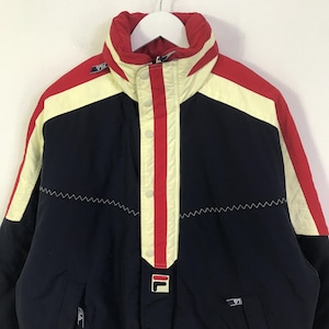 May include: A vintage Fila jacket in navy blue, red, and cream. The jacket features a high collar, a central red stripe with white buttons, and the Fila logo. The design includes a zigzag stitch detail and a white zipper.