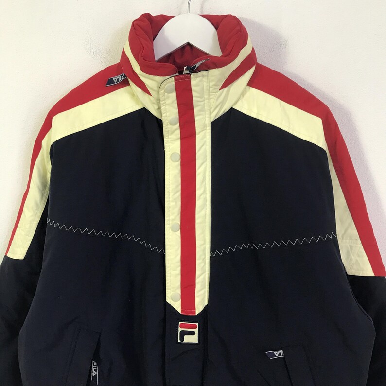 May include: A vintage Fila jacket in navy blue, red, and cream. The jacket features a high collar, a central red stripe with white buttons, and the Fila logo. The design includes a zigzag stitch detail and a white zipper.