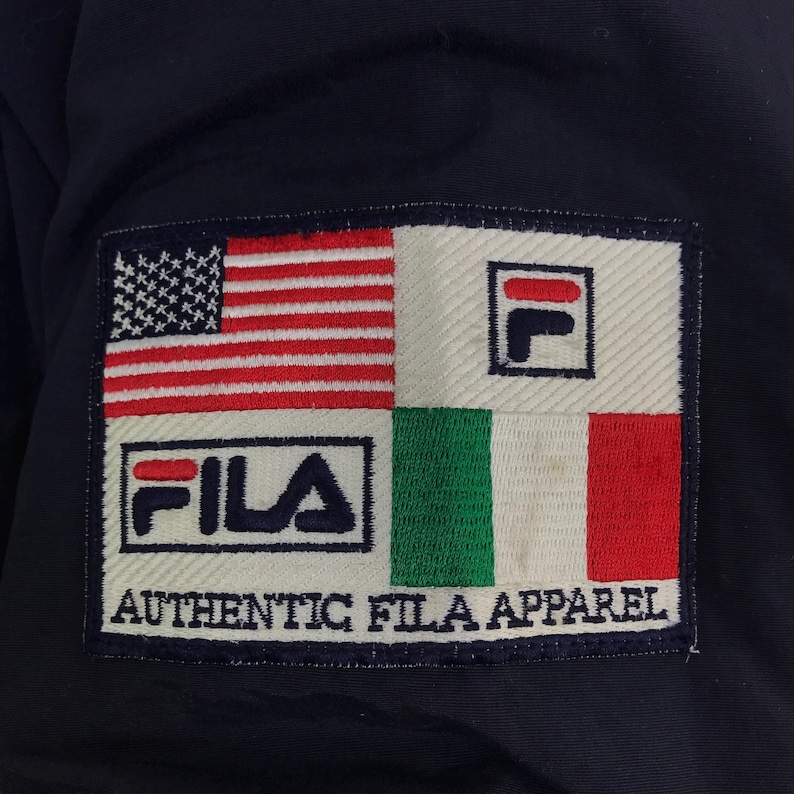 May include: Close-up of a navy blue jacket featuring a rectangular patch with the Fila logo and the text "AUTHENTIC FILA APPAREL." The patch incorporates the American and Italian flags, along with the Fila logo.