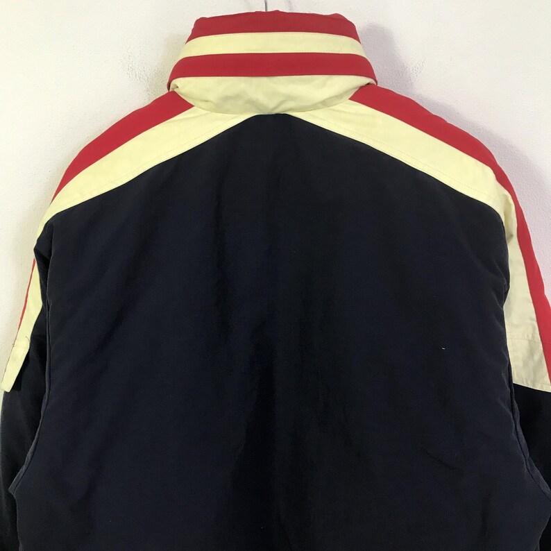 May include: A navy blue winter jacket with a high collar. The jacket features red and cream-colored stripes across the shoulders and collar. The back of the jacket is visible, showcasing the color-blocked design.