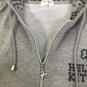 May include: A grey zip-up hoodie with a Hello Kitty design. The hood lining features a repeating pattern of Hello Kitty faces. The zipper has a star-shaped charm. The text "Hello Kitty" is printed on the front.