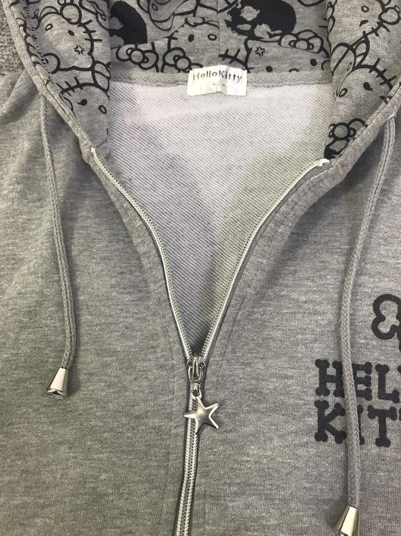 May include: A grey zip-up hoodie with a Hello Kitty design. The hood lining features a repeating pattern of Hello Kitty faces. The zipper has a star-shaped charm. The text "Hello Kitty" is printed on the front.