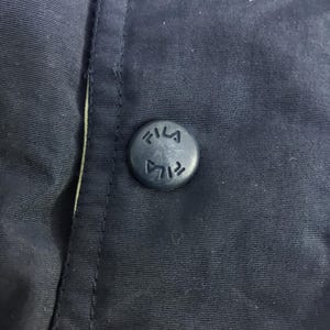 May include: Close-up of a navy blue button with the word "FILA" embossed on it. The button is attached to a navy blue fabric, with a seam visible on the left side. The button is circular and has a matte finish.