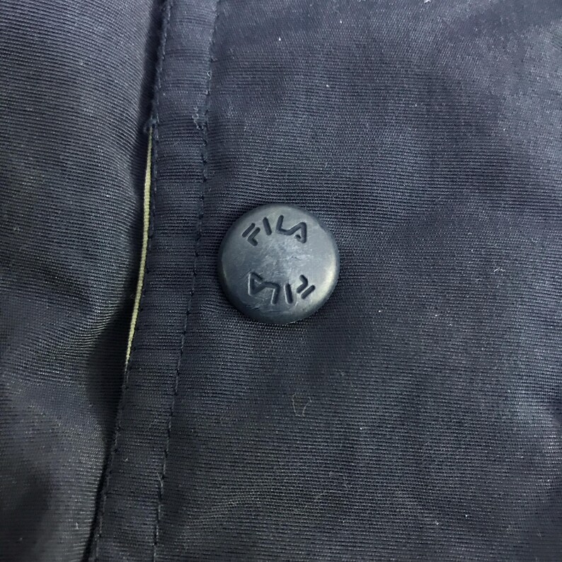 May include: Close-up of a navy blue button with the word "FILA" embossed on it. The button is attached to a navy blue fabric, with a seam visible on the left side. The button is circular and has a matte finish.